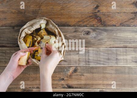 Die Hand der Frau legt Obstschalen in einen Papierbeutel auf Holzhintergrund. Recyclingkonzept. Stockfoto