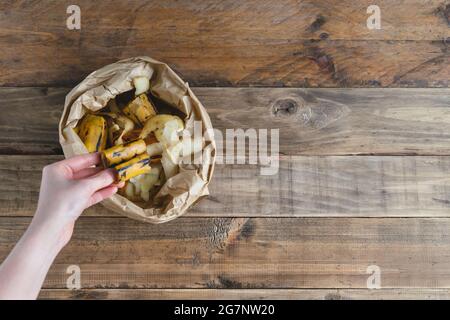 Frauenhand legt Bananenschale in Papiertüte auf Holzhintergrund ab. Speicherplatz kopieren. Stockfoto