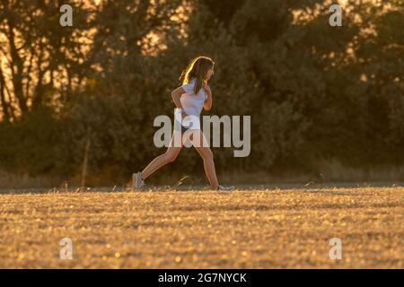 Teenagermädchen mit langen blonden Haaren, Shorts und weißem T-Shirt, das durch ein geerntetes Feld mit Bäumen im Hintergrund läuft. Sport im Fie Stockfoto