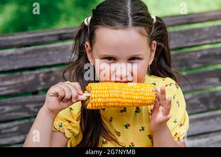 Ein charmantes sechsjähriges Mädchen mit Zöpfen in einem gelben Kleid hat ein köstliches Maiskolben auf einem Spaziergang zum Mittagessen Stockfoto