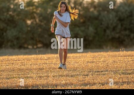 Teenager-Mädchen mit langen blonden Haaren, Shorts und weißem T-Shirt, das mit Blick auf die Kamera durch ein geerntetes Feld läuft. Sport auf dem Feld. Stockfoto