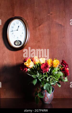 Wanduhr und frische rote und gelbe Rosen in Keramikkrug auf dunkelbraunem Holzhintergrund Stockfoto