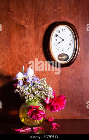 Wanduhr und verwelkte Blumen in Vase auf dunkelbraunem Holzhintergrund Stockfoto