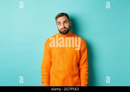 Trauriger und düsterer Mann schmollend, langweilen in der oberen linken Ecke, in orangefarbenem Pullover vor hellblauem Hintergrund stehend Stockfoto