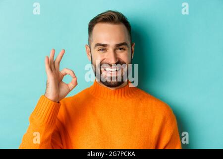 Nahaufnahme eines durchsetzungsfähigen jungen Mannes, der alles in Ordnung versicherte, ein Zeichen in Ordnung zeigte und lächelte, ja oder eine positive Antwort, auf hellblauem Hintergrund stehend Stockfoto