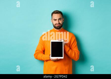 Technologie. Skeptisch und unzufrieden Kerl zeigt digitale Tablet-Bildschirm, runzelte enttäuscht, stand über hellblauen Hintergrund Stockfoto