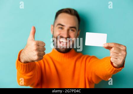 Nahaufnahme eines gutaussehenden kaukasischen Mannes, der auf einem türkisfarbenen Hintergrund zum Einkaufen geht, Kreditkarte und Daumen nach oben zeigt Stockfoto