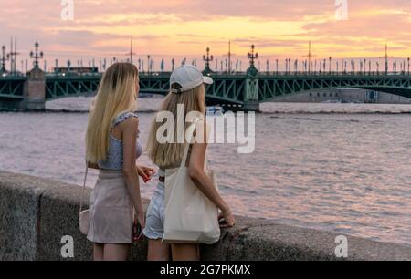 Zwei junge Frauen, die auf Granitdämmen stehen und sich lehnen und während der weißen Nacht in St. Petersburg den Sonnenuntergang über dem Fluss Neva genießen Stockfoto