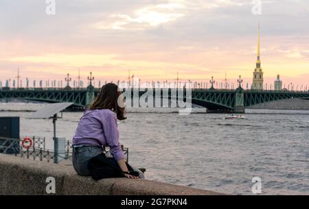 Junge Frau, die auf einem Granitdamm sitzt und den Sonnenuntergang hinter der Troitskiy-Brücke am Fluss Neva genießt, während der weißen Nacht, St. Petersburg, Russland Stockfoto