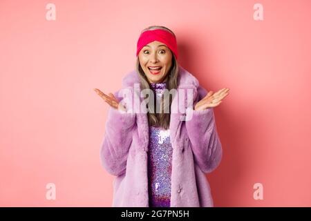 Mode- und Shopping-Konzept. Fröhliche asiatische ältere Frau in lila stilvollen Mantel überrascht, hören gute Nachrichten und jubeln, über rosa stehen Stockfoto