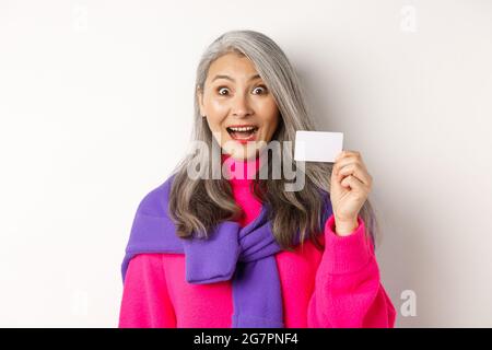 Einkaufskonzept. Glücklich asiatische alte aldy suchen beeindruckt und zeigt Kunststoff-Kreditkarte ihrer Bank, über weißem Hintergrund stehen Stockfoto