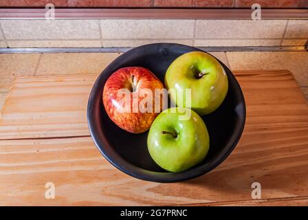 Nahaufnahme mehrerer reifer, reicher, frischer, organischer, saftiger roter und grüner Äpfel in einer schwarzen Keramikschale auf einem Holztisch. Kopierbereich.Selektiver Fokus. Stockfoto