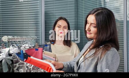 Geschäftsfrau mit langen lockeren Haaren wählt formelle Kleidung und Verkaufsassistent schlägt braune Bluse in der Modeboutique Nahansicht vor Stockfoto