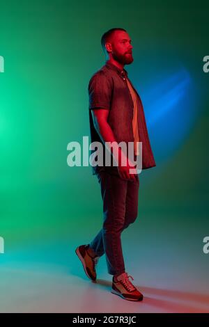 In voller Länge Studiobild des jungen kaukasischen bärtigen Mannes, der isoliert auf einem gradienten grün-blauen Hintergrund in Neonlichtern steht. Stockfoto