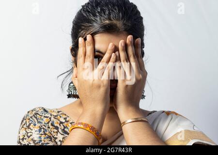 Isoliert auf weißem Hintergrund sieht ein indisches Weibchen vorsichtig aus, wobei ein Auge mit Händen auf weißem Hintergrund bedeckt ist Stockfoto
