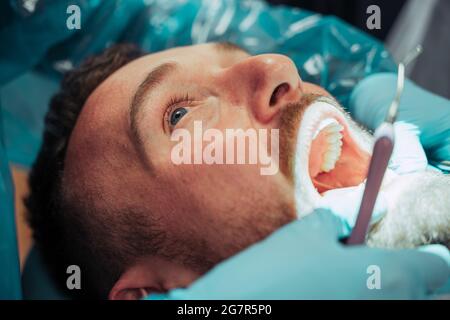 Kaukasischer Mann liegt mit weit geöffnetem Mund, während die männliche Krankenschwester an den Zähnen operiert Stockfoto