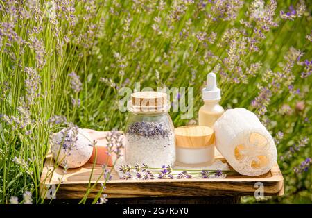 Verschiedene Beauty-Spa-Produkte auf Holztablett im blühenden Lavendelfeld am sonnigen Sommertag. Badesalz, Seifenstück, Tagescreme, Feuchtigkeitscreme, Badebombe. Stockfoto