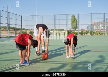 Basketballtrainer macht mit zwei Kindern Beindehnungs-Übungen, eines von ihnen hat eine Beinprothese. Trainer trainiert zwei Kinder. Stockfoto