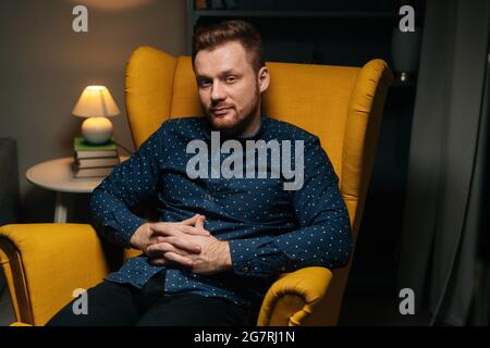 Mittellange Aufnahme eines selbstbewussten, gutaussehenden Mannes mit Bart, der ein stilvolles Hemd trägt und bequem in einem luxuriösen gelben Sessel sitzt. Stockfoto