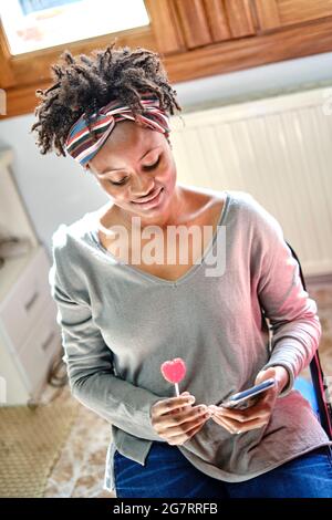 Nahaufnahme Porträt einer jungen schwarzen Frau mit afro Frisur scherzen mit einem Mobiltelefon und einem Lutscher innen zu Hause. Lifestyle-Konzept. Stockfoto