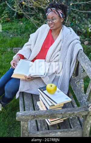 Porträt einer jungen schwarzen Frau mit Afro-Frisur und Brille, die ein Buch liest, das auf einer alten Gartenbank mit einem Bücherstapel sitzt. Lifestyle-Konzept. Stockfoto
