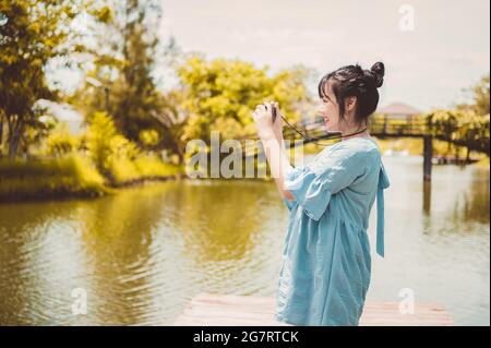 Asiatische Frau in blauem Kleid im öffentlichen Park trägt digitale spiegellose Kamera und macht Foto ohne Gesichtsmaske in glücklicher Stimmung. Menschen Lebensstil und l Stockfoto