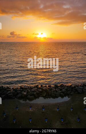 Vertikales Panorama des schönen Sonnenuntergangs über dem Meer und Strand voller Sonnenschirme mit Sonne hinter Wolken Stockfoto