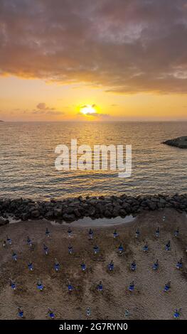 Vertikales Panorama des schönen Sonnenuntergangs über dem Meer und Strand voller Sonnenschirme mit Sonne hinter Wolken Stockfoto