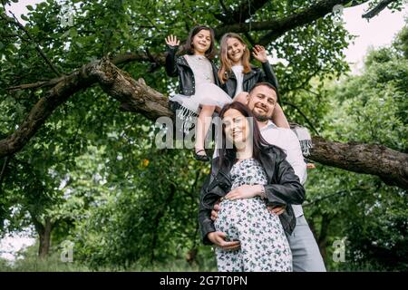 Attraktive Schwangerin mit ihrem Mann und zwei Töchtern, die an der frischen Luft posieren. Glückliche Familie umarmen und lächeln während der Zeit im Freien verbringen. Stockfoto