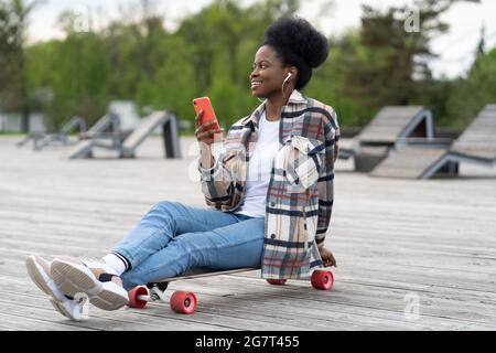 Happy african Mädchen halten Smartphone sitzen im städtischen Park auf Longboard entspannt Musik hören und Messaging Stockfoto