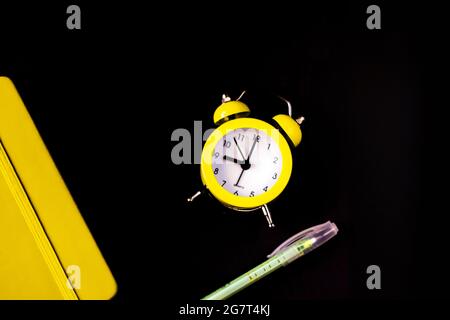 Direkt oberhalb Aufnahme von gelbem Wecker mit geschlossenem Notizblock Tagebuch auf schwarzem Hintergrund. Zeitmanagement-Konzept. Zurück zur Schule. Selektiver Fokus Stockfoto