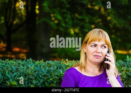 Unschärfe kaukasisch blonde Frau reden, sprechen am Telefon draußen, im Freien. 40-jährige Frau in lila Bluse im Park auf der Bank. Erwachsene Person usi Stockfoto