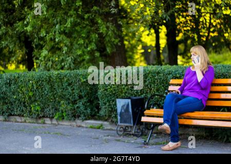 Unschärfe-Eleganz kaukasische blonde Frau reden, sprechen auf dem Telefon draußen, im Freien. 40-jährige Frau in lila Bluse im Park. Erwachsene Frauen usin Stockfoto