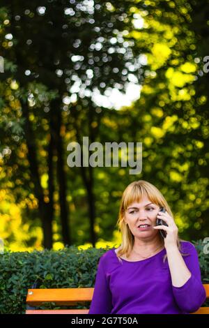 Unschärfe kaukasisch blonde Frau reden, sprechen am Telefon draußen, im Freien. 40-jährige Frau in lila Bluse im Park. Erwachsene Personen, die ein Mobiltelefon verwenden Stockfoto