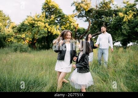 Zwei fröhliche Schwestern spielen zusammen im grünen Garten, während ihre Eltern die Hände halten und auf dem Hintergrund laufen. Fröhliche vierköpfige Familie, die ihre Freizeit an der frischen Luft genießen kann. Stockfoto