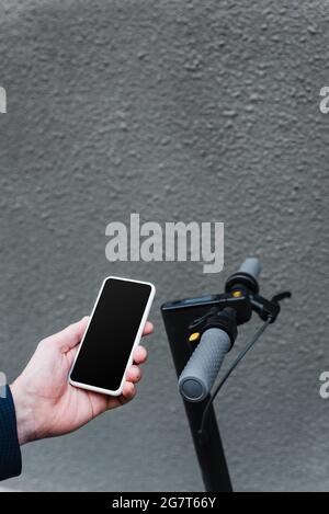 Teilansicht des reifen Geschäftsmannes, der Smartphone mit leerem Bildschirm in der Nähe des E-Scooters hält Stockfoto