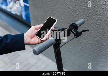 Beschnittene Ansicht eines reifen Geschäftsmannes mit Smartphone und leerem Bildschirm in der Nähe eines E-Scooters Stockfoto