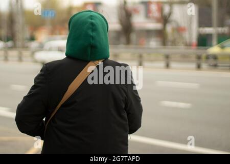 Der Mann in der Haube. Ein unbekannter Mann auf der Straße. Ein freundlich gekleideter Bürger. Grüne Kapuze und schwarze Jacke. Den Gürtel über seiner Schulter. Stockfoto