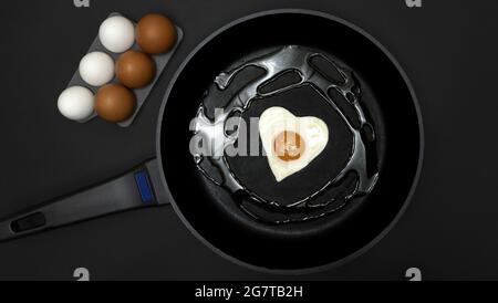 Eine Pfanne mit einem Spiegelei in Herzform und daneben ein halbes Dutzend weißer und brauner Eier, alles auf schwarzem Hintergrund, gesundes Kochen und Stockfoto