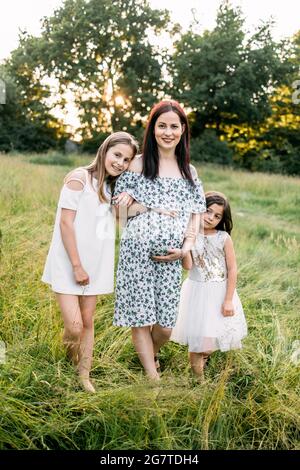 Schöne Schwangere mit zwei niedlichen Töchtern, die Freizeit in der Natur verbringen. Kleine Schwestern stehen in der Nähe der Mutter und umarmen den schwanger Bauch. Liebe und Familie Konzept. Stockfoto