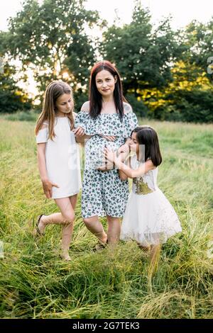 Schöne Schwangere mit zwei niedlichen Töchtern, die Freizeit in der Natur verbringen. Kleine Schwestern stehen in der Nähe der Mutter und umarmen den schwanger Bauch. Liebe und Familie Konzept. Stockfoto