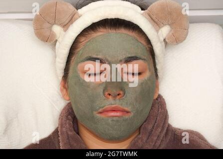 Hübsche junge Frau mit blauer Tonmaske zu Hause. Reinigungsverfahren Maske für fettige Akne-peigende Problemhaut. Dermatologische häusliche Verfahren. Stockfoto