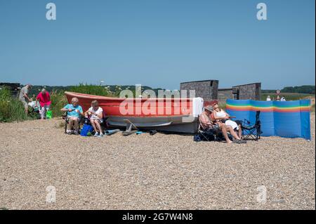 Blick auf ein rotes Boot am Kiesstrand mit Leuten, die vor dem Hotel sitzen und Fisch und Chips vom lokalen Restaurant in Dunwich Suffolk essen Stockfoto