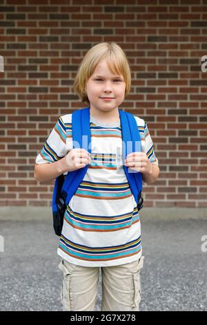 Schönes lächelndes Kind, das in der Nähe des Schulgebäudes auf dem Schulhof gegen eine Ziegelmauer steht. Kleine blonde Student trägt blauen Rucksack. Zurück zur Schule con Stockfoto