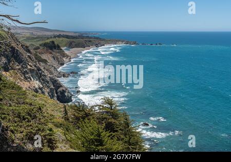 San Simeon, CA, USA - 8. Juni 2021: Pazifikküste. Weiße Brandung des blauen Ozeans stürzt gegen schwarze Felsen ab. Weiter unten befindet sich ein Sandstrand, Gree Stockfoto