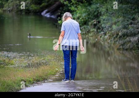 Hattingen, NRW, Deutschland. Juli 2021. Eine Frau geht auf einem normalerweise beliebten Pfad entlang des Flusses, der jetzt im Wasser untergetaucht ist. Die Ruhr hat ihre Böschung, Felder und viele Gärten, Keller und Grundstücke in der Nähe der Stadt Hattingen im Ruhrgebiet in Nordrhein-Westfalen überflutet. NRW ist nach starken Regenfällen in den letzten Tagen von schrecklichen Überschwemmungen heimgesucht worden. Bei den Überschwemmungen in Deutschland sind bisher mehr als 80 Menschen ums Leben gekommen. Kredit: Imageplotter/Alamy Live Nachrichten Stockfoto