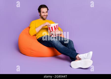Portrait von attraktiven fröhlichen Mann sitzt in Tasche Stuhl mit Spaß essen Mais Fernsehen isoliert über pastellviolett lila Farbe Hintergrund Stockfoto