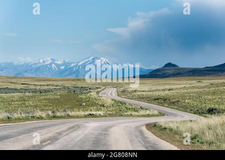 Kurvenreiche Straße in Richtung der schneebedeckten Berge am Horizont mit einigen Prärien um sie herum, im Antelope Island State Park, in der Nähe von Salt Lake City, Ut Stockfoto