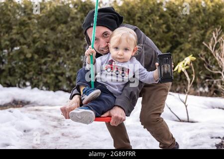 Kanadischer Vater und blonder Baby-Sohn posieren für die Kamera in einem verschneiten Spätwintergarten und lächeln der Kamera zu, während sie mit einer Schaukel spielen. Stockfoto