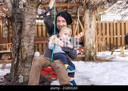 Der kanadische Vater, sein Kleinkind, posiert in einem verschneiten Spätwintergarten für die Kamera und lächelt dabei mit einer Gartenschaukel vor der Kamera. Stockfoto